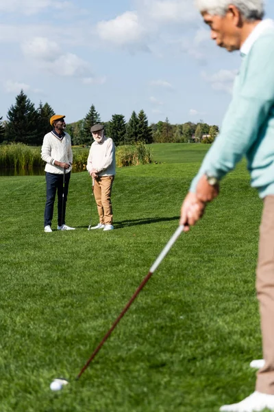 Sênior interracial homens no apartamento bonés olhando para asiático amigo jogar golfe no embaçado primeiro plano — Fotografia de Stock