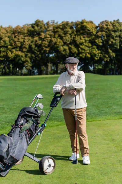 Bearded senior man in flat cap standing near golf cart — стоковое фото