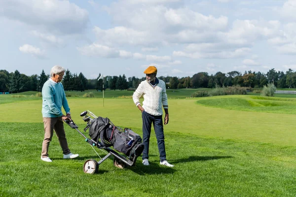 Asiático sénior hombre caminando con golf carro cerca rico africano americano amigo - foto de stock