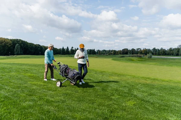 Sênior asiático homem andando com golfe carrinho perto rico africano americano amigo — Fotografia de Stock