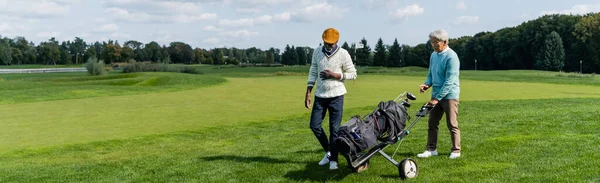 Ásia sênior homem andando com golfe carrinho perto rico africano americano amigo, banner — Fotografia de Stock