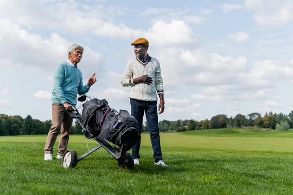 Asian senior man pointing away while walking with golf cart near wealthy african american friend — стоковое фото