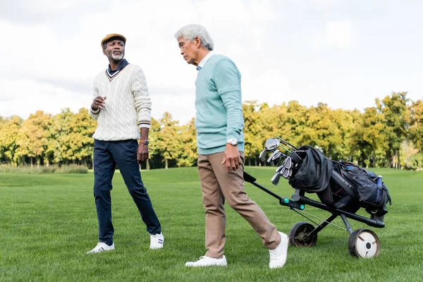 Africano americano hombre caminando y hablando con senior asiático amigo cerca golf carro - foto de stock