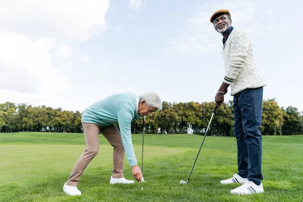 Asiático senior hombre poniendo bola en golf tee cerca africano americano amigo - foto de stock
