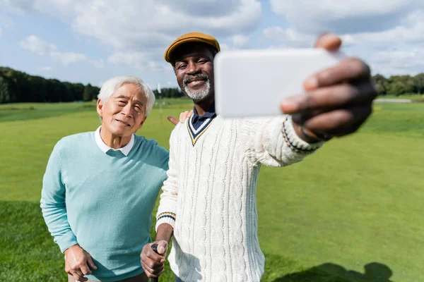 Feliz africano americano hombre tomando selfie con senior asiático amigo en borrosa smartphone - foto de stock