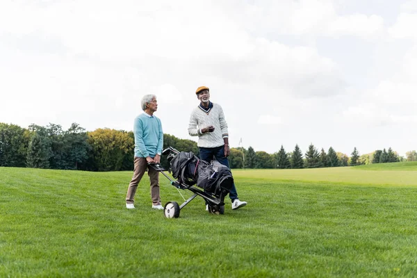 Asiático senior hombre caminando con golf carro cerca africano americano amigo - foto de stock
