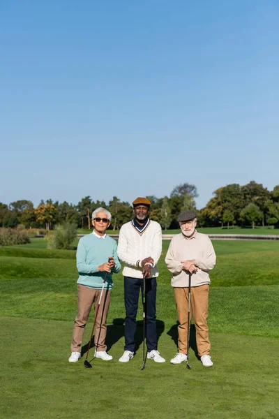 Asian man in sunglasses standing near senior multiethnic friends in flat caps with golf clubs — Stock Photo