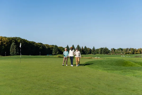 Senior asian man in sunglasses walking near interracial friends with golf clubs — Stock Photo