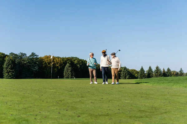 Senior asian man in sunglasses walking near multiethnic friends with golf clubs — стоковое фото