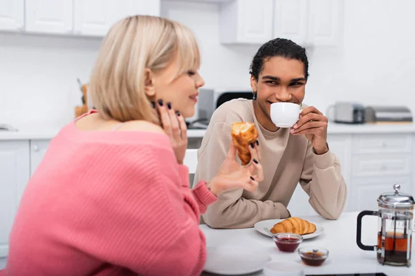 Happy african american man drinking tea and looking at girlfriend with croissant — стоковое фото