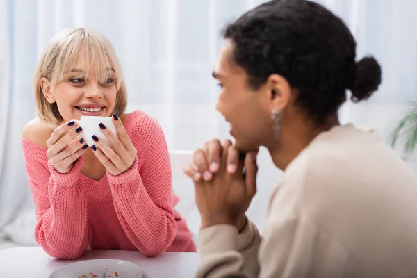 Happy blonde woman holding cup and looking at african american man — Stock Photo