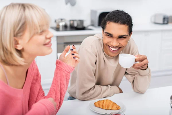 Smiling african american man holding cup of tea and looking at blurred blonde girlfriend — Stock Photo