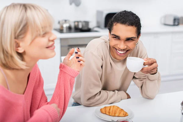 Smiling african american man holding cup of tea and looking at blurred blonde girlfriend — стоковое фото