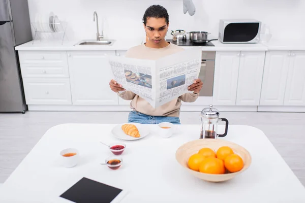 Junger afrikanisch-amerikanischer Mann liest Zeitung in der Nähe des Frühstücks und digitales Tablet auf dem Tisch — Stockfoto