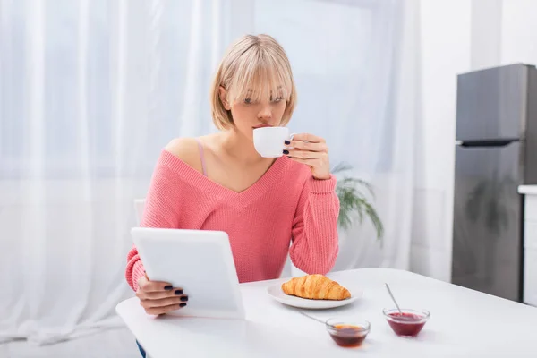 Молода блондинка п'є чай і використовує цифровий планшет під час сніданку — стокове фото