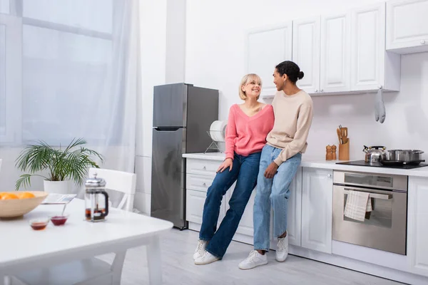 Full length of happy multiethnic couple looking at each other in modern kitchen — Fotografia de Stock