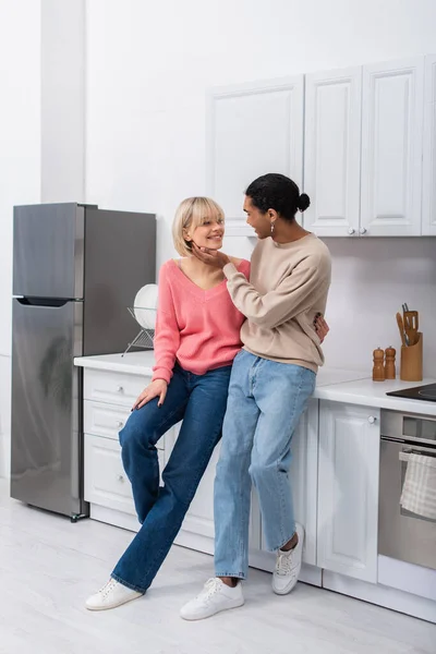 Full length of smiling multiethnic couple looking at each other in modern kitchen — Foto stock