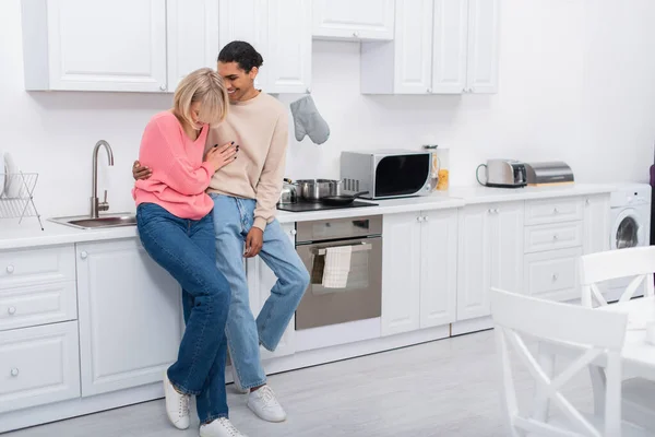 Full length of pleased multiethnic couple standing in modern kitchen — стоковое фото