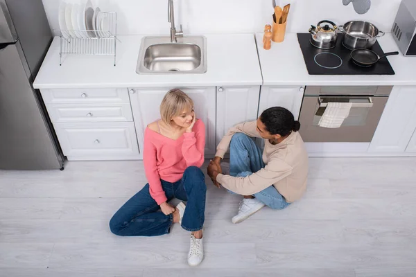 Vista superior de casal multiétnico feliz sentado no chão na cozinha moderna — Fotografia de Stock