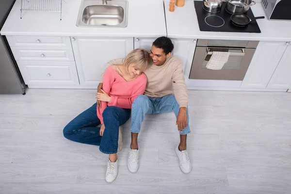 Vista superior de alegre casal multiétnico sentado no chão na cozinha moderna — Fotografia de Stock