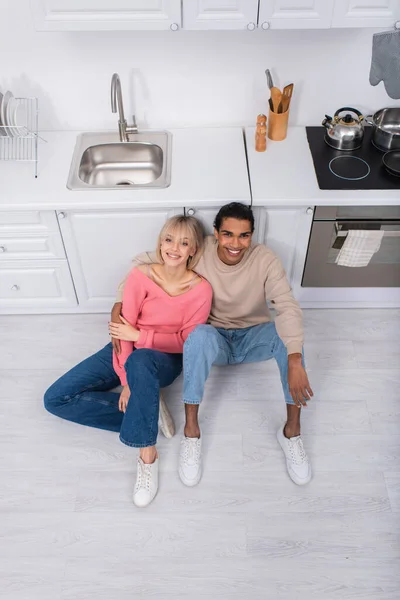 Vista superior de pareja multiétnica positiva sentada en el suelo en la cocina moderna - foto de stock