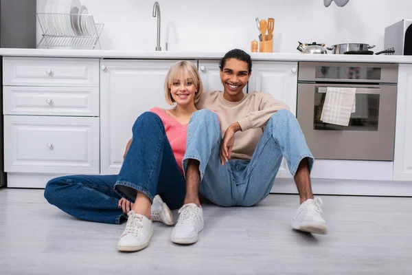 Positives multiethnisches Paar sitzt in moderner Küche auf dem Boden — Stockfoto