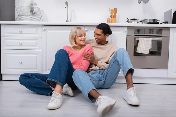 Alegre casal multiétnico sentado no chão na cozinha moderna — Fotografia de Stock