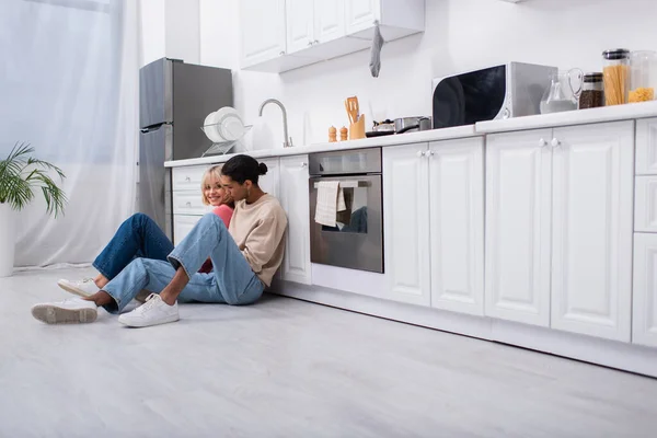 Glückliches junges multiethnisches Paar in moderner Küche auf dem Boden sitzend — Stockfoto