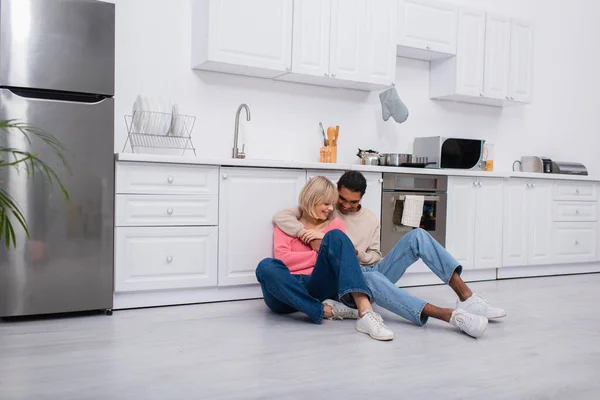 Позитивна багатоетнічна пара обіймає і сидить на підлозі в сучасній кухні — стокове фото