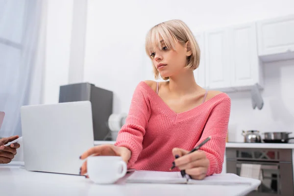 Young blonde woman reaching cup while writing on notebook near laptop — Stock Photo