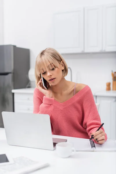 Joven freelancer hablando en el teléfono inteligente mientras escribe cerca del ordenador portátil - foto de stock