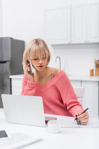 Mujer rubia joven hablando en el teléfono inteligente mientras escribe cerca del ordenador portátil - foto de stock