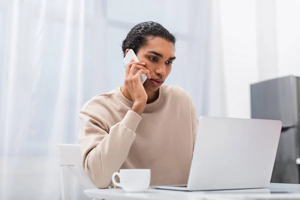 Joven afroamericano freelancer hablando en smartphone cerca de portátil y taza - foto de stock