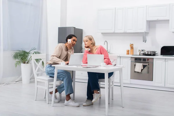 Young african american freelancer talking on smartphone near blonde girlfriend and laptops — Stockfoto