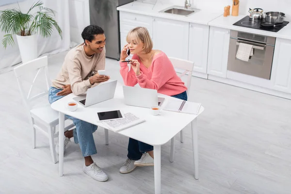 Vista de ángulo alto del hombre afroamericano feliz mirando a la novia rubia hablando en el teléfono inteligente mientras trabaja desde casa - foto de stock