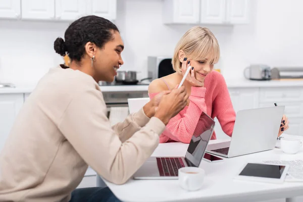 Glücklicher afrikanisch-amerikanischer Mann mit Smartphone in der Nähe lächelnder Freundin, die von zu Hause aus am Laptop arbeitet — Stockfoto