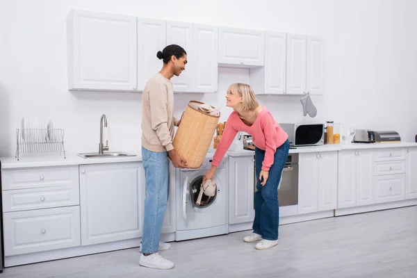 Happy blonde woman putting clothes in washing machine near african american man — Stock Photo