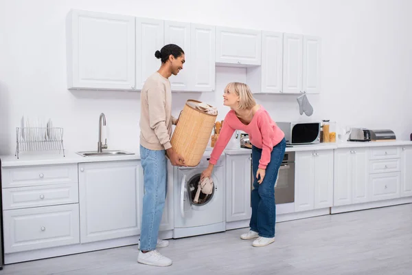 Happy blonde woman putting clothes in washing machine near african american man - foto de stock