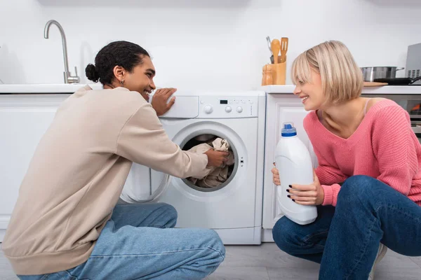 Felice donna bionda in possesso di bottiglia con detergente vicino afro-americano uomo mettere i vestiti in lavatrice — Foto stock