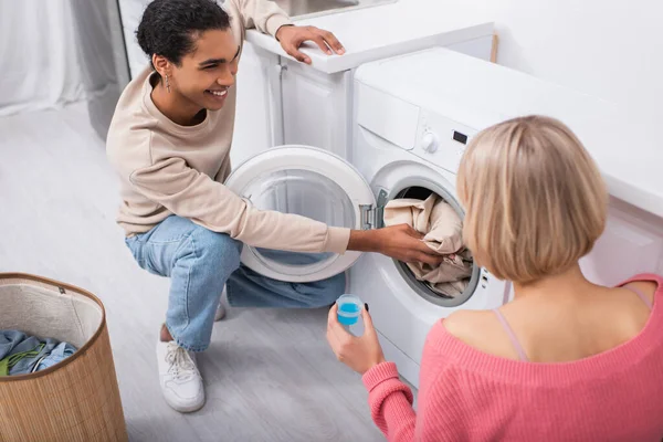 Felice donna bionda in possesso di tazza con detergente vicino uomo americano africano mettere i vestiti in lavatrice — Foto stock