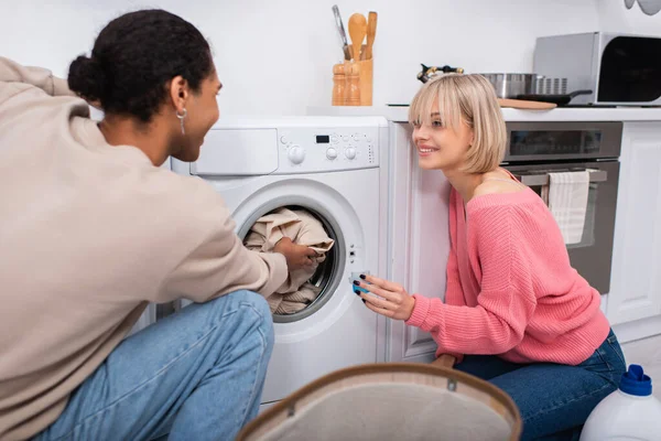 Gaie femme blonde tenant tasse avec détergent près de l'homme afro-américain mettre des vêtements dans la machine à laver — Photo de stock