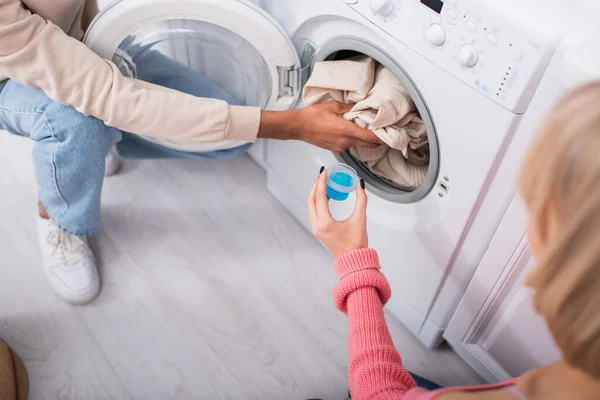 Vista ritagliata della donna che tiene tazza con detergente vicino all'uomo americano africano che mette i vestiti in lavatrice — Foto stock