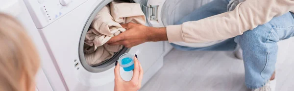 Vista ritagliata della donna che tiene la tazza con detergente vicino all'uomo americano africano che mette i vestiti in lavatrice, banner — Foto stock
