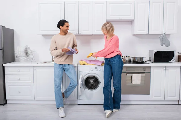 Cheerful interracial couple holding clean and colorful clothes — стоковое фото