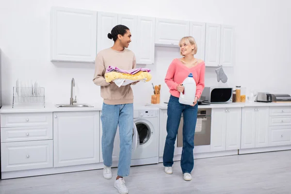 Full length of happy interracial couple holding bottle with detergent and colorful clothes — Stock Photo