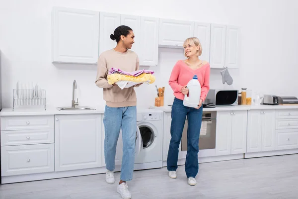 Pleine longueur de couple interracial heureux tenant bouteille avec du détergent et des vêtements colorés — Photo de stock