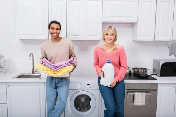 Heureux couple interracial tenant bouteille avec détergent et vêtements colorés — Photo de stock