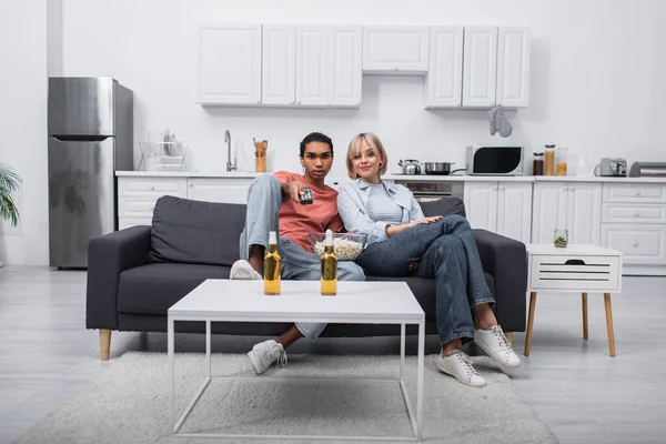 Afrikanisch-amerikanischer Mann hält Fernbedienung in der Hand und klickt Kanäle in der Nähe der blonden Freundin im Wohnzimmer — Stockfoto
