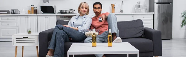 Africano americano homem segurando controlador remoto e clicando canais perto de namorada loira na sala de estar, banner — Fotografia de Stock