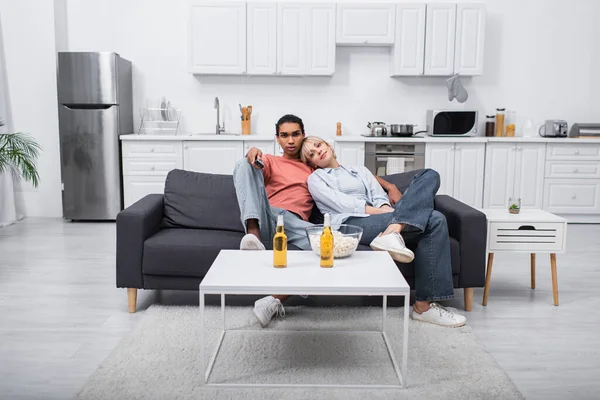 Young blonde woman leaning on african american man holding remote controller in living room — Fotografia de Stock